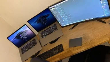 A wooden desk with two laptops, a curved monitor, keyboard and trackpad